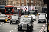 A busy urban street scene featuring iconic black London taxis and red double-decker buses among various other vehicles. Large buildings with intricate architecture rise in the background, and a prominent billboard features an advertisement. The traffic seems to be organized yet congested.