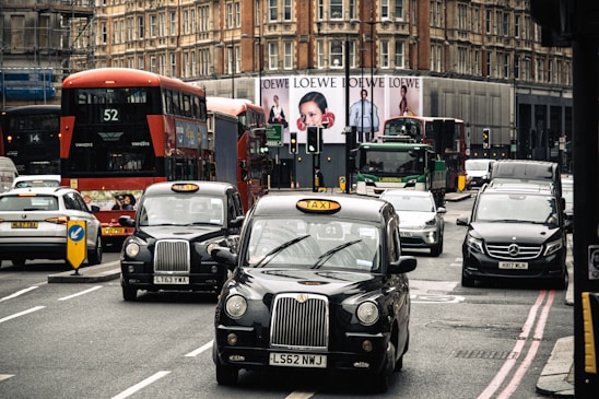 A busy urban street scene featuring iconic black London taxis and red double-decker buses among various other vehicles. Large buildings with intricate architecture rise in the background, and a prominent billboard features an advertisement. The traffic seems to be organized yet congested.
