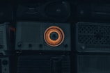 A collection of vintage radios with a prominent one in the center featuring an illuminated orange dial. The overall scene is dimly lit, emphasizing the nostalgic and retro aesthetic of the objects.