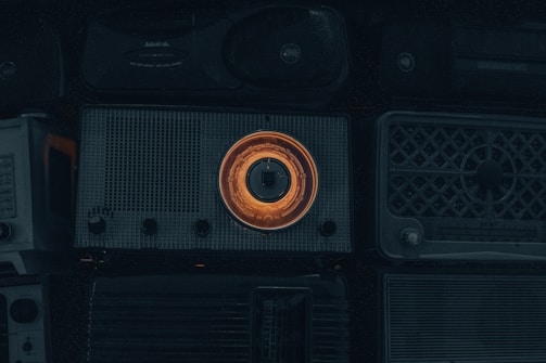 A collection of vintage radios with a prominent one in the center featuring an illuminated orange dial. The overall scene is dimly lit, emphasizing the nostalgic and retro aesthetic of the objects.