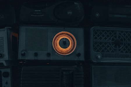 A collection of vintage radios with a prominent one in the center featuring an illuminated orange dial. The overall scene is dimly lit, emphasizing the nostalgic and retro aesthetic of the objects.