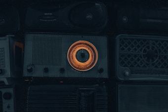 A collection of vintage radios with a prominent one in the center featuring an illuminated orange dial. The overall scene is dimly lit, emphasizing the nostalgic and retro aesthetic of the objects.