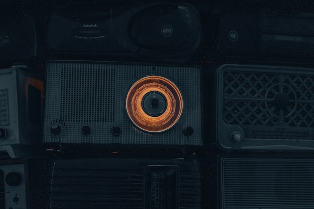 A collection of vintage radios with a prominent one in the center featuring an illuminated orange dial. The overall scene is dimly lit, emphasizing the nostalgic and retro aesthetic of the objects.