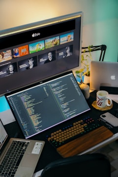 A workspace with a multi-monitor setup featuring a coding environment on the main screen and Apple TV+ content on a secondary screen. The desk includes a keyboard, laptop, a mug, and a plant. The atmosphere is tech-focused and organized, illuminated by ambient lighting.