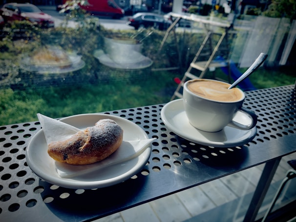 A cup of coffee with latte art sits on a saucer next to a small, sugar-dusted pastry on a metal table with a perforated pattern. The setting is by a window, showing an outdoor area with greenery and parked cars.