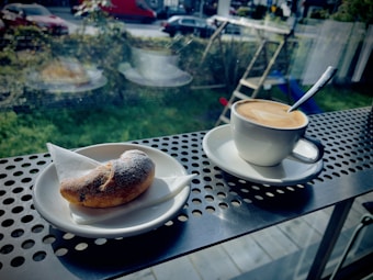 A cup of coffee with latte art sits on a saucer next to a small, sugar-dusted pastry on a metal table with a perforated pattern. The setting is by a window, showing an outdoor area with greenery and parked cars.