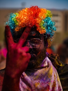 A person with a colorful curly wig featuring orange, blue, green, and yellow hues. The face is covered in colored powders, suggestive of a festive celebration, and they are wearing reflective sunglasses. The person is making a peace sign gesture with their fingers.