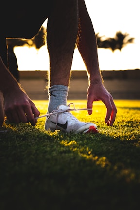 Athlete tying shoelaces of high-performance black and red cleats on a vibrant green field at dusk.