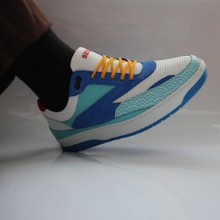 A colorful sneaker featuring a combination of blue, white, turquoise, and yellow colors. The shoe has a unique design with wave patterns on the side and bright yellow laces. The sneaker is worn with dark socks and brown pants, set against a neutral background.