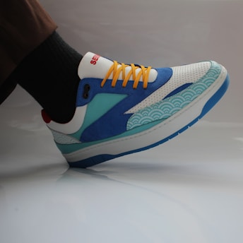A colorful sneaker featuring a combination of blue, white, turquoise, and yellow colors. The shoe has a unique design with wave patterns on the side and bright yellow laces. The sneaker is worn with dark socks and brown pants, set against a neutral background.
