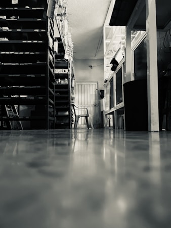 The image portrays a room with reflective flooring, shelves filled with various items on either side, and a single plastic chair positioned in the middle near a doorway. The setting is likely a storage or utility space.