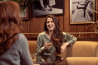 Two people are engaged in a conversation at a cozy café, with a woman in focus smiling and holding a drink. The setting includes warm wooden walls and framed black-and-white photographs in the background.