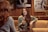 Two people are engaged in a conversation at a cozy café, with a woman in focus smiling and holding a drink. The setting includes warm wooden walls and framed black-and-white photographs in the background.