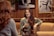Two people are engaged in a conversation at a cozy café, with a woman in focus smiling and holding a drink. The setting includes warm wooden walls and framed black-and-white photographs in the background.