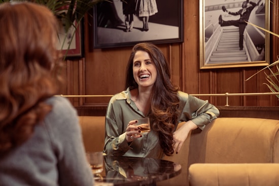 Two people are engaged in a conversation at a cozy caf&eacute;, with a woman in focus smiling and holding a drink. The setting includes warm wooden walls and framed black-and-white photographs in the background.