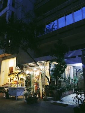 A dimly lit floral shop nestled between urban buildings in the evening, with a glowing display of flower arrangements under a canopy decorated with hanging lights. Potted plants are scattered around the entrance, adding a touch of nature to the concrete surroundings.