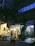 A dimly lit floral shop nestled between urban buildings in the evening, with a glowing display of flower arrangements under a canopy decorated with hanging lights. Potted plants are scattered around the entrance, adding a touch of nature to the concrete surroundings.