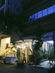 A dimly lit floral shop nestled between urban buildings in the evening, with a glowing display of flower arrangements under a canopy decorated with hanging lights. Potted plants are scattered around the entrance, adding a touch of nature to the concrete surroundings.
