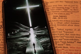 An open book featuring text in a contrasting color to the background, with a smartphone placed over it. The phone displays an image of a person walking down a path towards a bright, glowing cross set against a dark landscape.