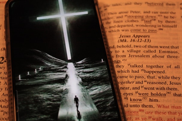 An open book featuring text in a contrasting color to the background, with a smartphone placed over it. The phone displays an image of a person walking down a path towards a bright, glowing cross set against a dark landscape.