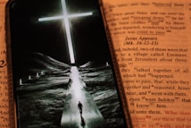 An open book featuring text in a contrasting color to the background, with a smartphone placed over it. The phone displays an image of a person walking down a path towards a bright, glowing cross set against a dark landscape.