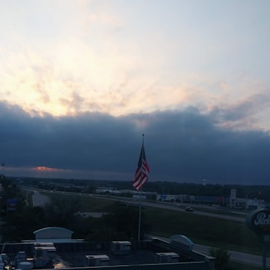 An American flag subtly waving behind a rugged semi-truck on a highway at sunset.