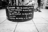 A black and white image of a large cylindrical planter sitting on a tiled pavement. The planter has a poem written in Portuguese painted on its side. In the background, a person wearing shorts and sneakers walks past a public square or open space with buildings and other people.
