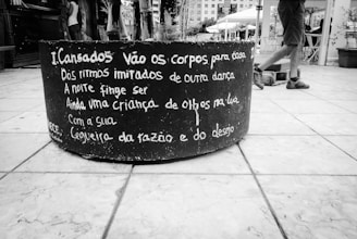 A black and white image of a large cylindrical planter sitting on a tiled pavement. The planter has a poem written in Portuguese painted on its side. In the background, a person wearing shorts and sneakers walks past a public square or open space with buildings and other people.