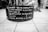 A black and white image of a large cylindrical planter sitting on a tiled pavement. The planter has a poem written in Portuguese painted on its side. In the background, a person wearing shorts and sneakers walks past a public square or open space with buildings and other people.
