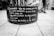 A black and white image of a large cylindrical planter sitting on a tiled pavement. The planter has a poem written in Portuguese painted on its side. In the background, a person wearing shorts and sneakers walks past a public square or open space with buildings and other people.