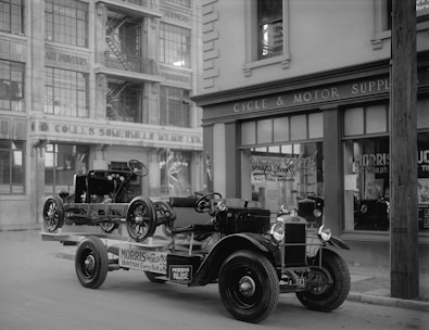 A vintage black and white photograph of an early 20th-century street scene featuring a classic Morris truck carrying a car chassis. The background shows a multi-story building with signs for art printers and stationers. There is also a storefront labeled 'Cycle & Motor Supply'. The street and architecture suggest an urban setting.