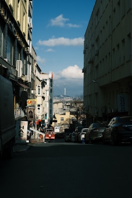 A narrow street lined with buildings on both sides, leading towards a distant cityscape. The road is populated with parked cars and a few signs in a foreign language. In the background, a tall tower is visible against a partly cloudy sky, suggesting a blend of urban life and architectural elements.