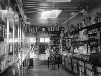 A vintage store interior displaying shelves packed with various bottles and boxes. Glass cabinets along the counters showcase neatly arranged products. A prominent clock sits above the shelves, and an empty chair is positioned in the middle of the floor. The store has a high ceiling with light coming in through skylights.