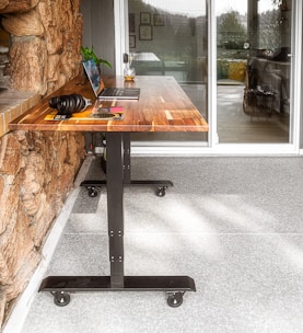 A wooden table with metal legs on wheels is placed against a stone wall. A laptop and a pair of headphones rest on the table, along with a book and a plant. A glass of what appears to be a beverage is also on the table. In the background, there is a large glass door leading to an indoor space.