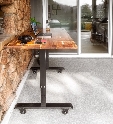 A wooden table with metal legs on wheels is placed against a stone wall. A laptop and a pair of headphones rest on the table, along with a book and a plant. A glass of what appears to be a beverage is also on the table. In the background, there is a large glass door leading to an indoor space.
