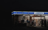 A brightly lit convenience store at night with large glass windows and visible interior displays of various products. Several people are seen inside and near the entrance, suggesting activity and commerce.