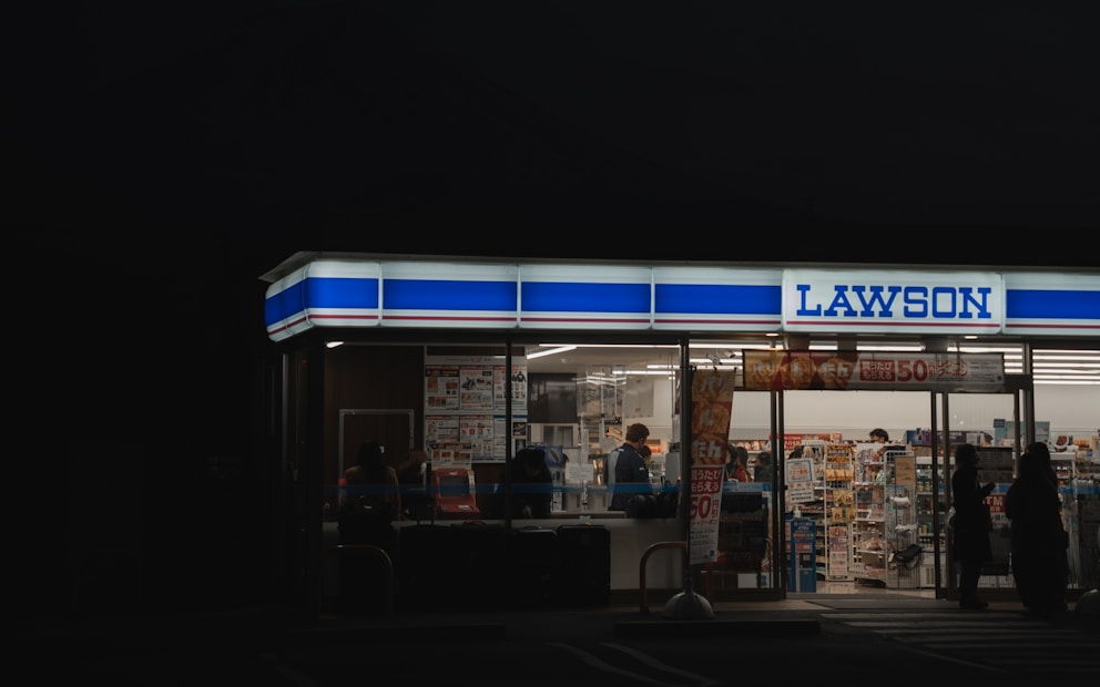 A brightly lit convenience store at night with large glass windows and visible interior displays of various products. Several people are seen inside and near the entrance, suggesting activity and commerce.