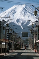 A street lined with traditional Japanese signs and lamp posts, with Mount Fuji towering majestically in the background. The street is mostly empty, with a person walking towards the center and a vehicle further down.