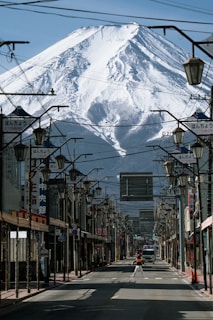 A street lined with traditional Japanese signs and lamp posts, with Mount Fuji towering majestically in the background. The street is mostly empty, with a person walking towards the center and a vehicle further down.