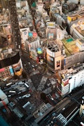 A bustling urban intersection surrounded by numerous tall buildings adorned with colorful billboards and advertisements. The streets below are filled with sparse crowds of people and a few vehicles. The scene is vibrant with commercial activity and dense infrastructure.