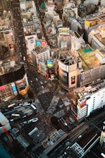 A bustling urban intersection surrounded by numerous tall buildings adorned with colorful billboards and advertisements. The streets below are filled with sparse crowds of people and a few vehicles. The scene is vibrant with commercial activity and dense infrastructure.