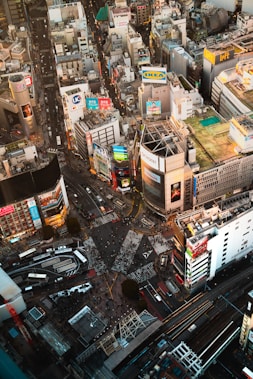 A bustling urban intersection surrounded by numerous tall buildings adorned with colorful billboards and advertisements. The streets below are filled with sparse crowds of people and a few vehicles. The scene is vibrant with commercial activity and dense infrastructure.