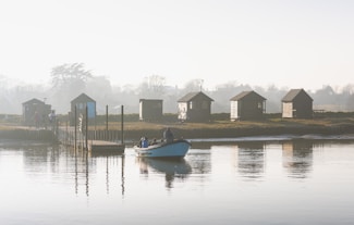 Several small wooden cabins are situated along a tranquil riverside, with a dock extending into the water. A small boat carrying a few people is approaching the dock. The air appears misty, creating a serene and calm atmosphere with soft light filtering through.