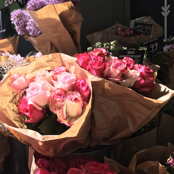 Bouquets of pink and red roses wrapped in brown paper are displayed in a floral shop. The roses are vibrant, with a mix of tightly closed buds and blooming flowers. In the background, there are other flowers such as purple blooms, and price tags are visible.