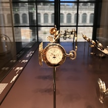 An ornate, antique pocket watch with intricate detailing is prominently displayed on a stand. It features a set of dials and Roman numerals, surrounded by other similar vintage timepieces, all showcased in a well-lit glass case. The background shows a large window with views of an architectural structure.