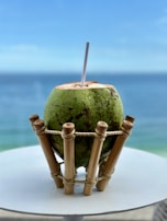 A fresh green coconut placed on a bamboo holder with a straw inserted at the top. It is set on a white surface, against a background of a calm blue ocean under a clear sky.