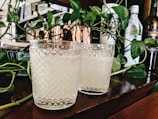 Two textured glass tumblers filled with a light beverage are placed on a wooden surface. They are surrounded by green leafy vines and bottles in the background, creating a cozy and natural setting.