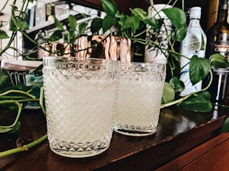 Two textured glass tumblers filled with a light beverage are placed on a wooden surface. They are surrounded by green leafy vines and bottles in the background, creating a cozy and natural setting.