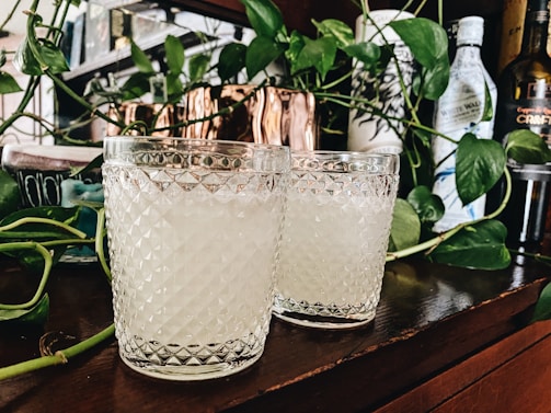 Two textured glass tumblers filled with a light beverage are placed on a wooden surface. They are surrounded by green leafy vines and bottles in the background, creating a cozy and natural setting.