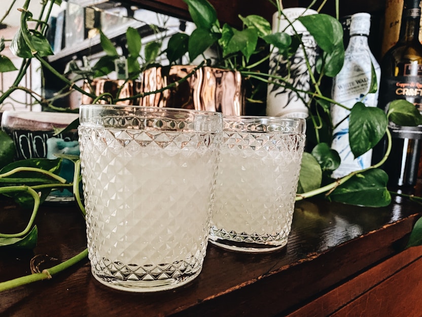 Two textured glass tumblers filled with a light beverage are placed on a wooden surface. They are surrounded by green leafy vines and bottles in the background, creating a cozy and natural setting.